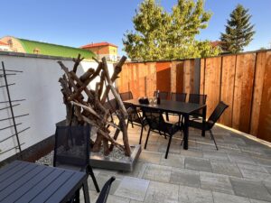 Venkovní posezení na dlážděné terase s černým stolem, židlemi a dekorativní dřevěnou plastikou.