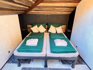 Manželská postel se zeleným ložním prádlem v ložnici s dřevěným stropem a tmavou stěnou za hlavou.