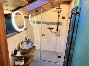 Koupelna v podkroví s kamenným umyvadlem na dřevěné desce, kulatým podsvíceným zrcadlem a proskleným sprchovým koutem.