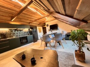 Pohled na jídelní stůl s modrými židlemi a tmavou kuchyňskou linku pod šikmým dřevěným stropem.